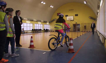 Element dekoracyjny - uczennica na rowerze pokonuje slalomem tor przeskód zlokalizowany w hali spotrowej